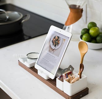 A kitchen counter with a wooden organizer holding a tablet displaying a recipe, along with small kitchen tools, with a bowl of limes in the background.