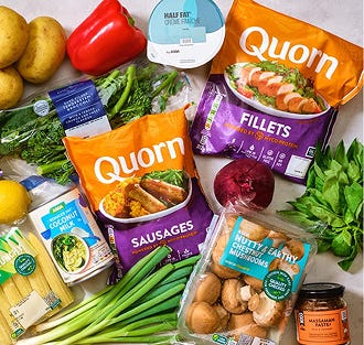 A variety of fresh vegetables and Quorn products, including pieces, sausages, and fillets, arranged on a light surface.
