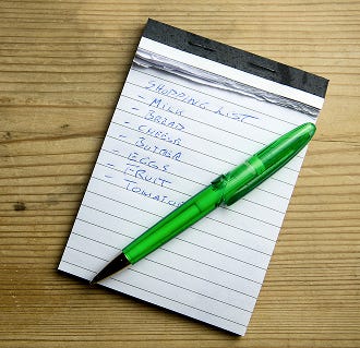 A green pen on a notepad with a handwritten shopping list on a wooden surface.