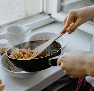 A person is using a wooden spatula to stir food in a black frying pan, with steam rising from the pan, next to a sunlit window.